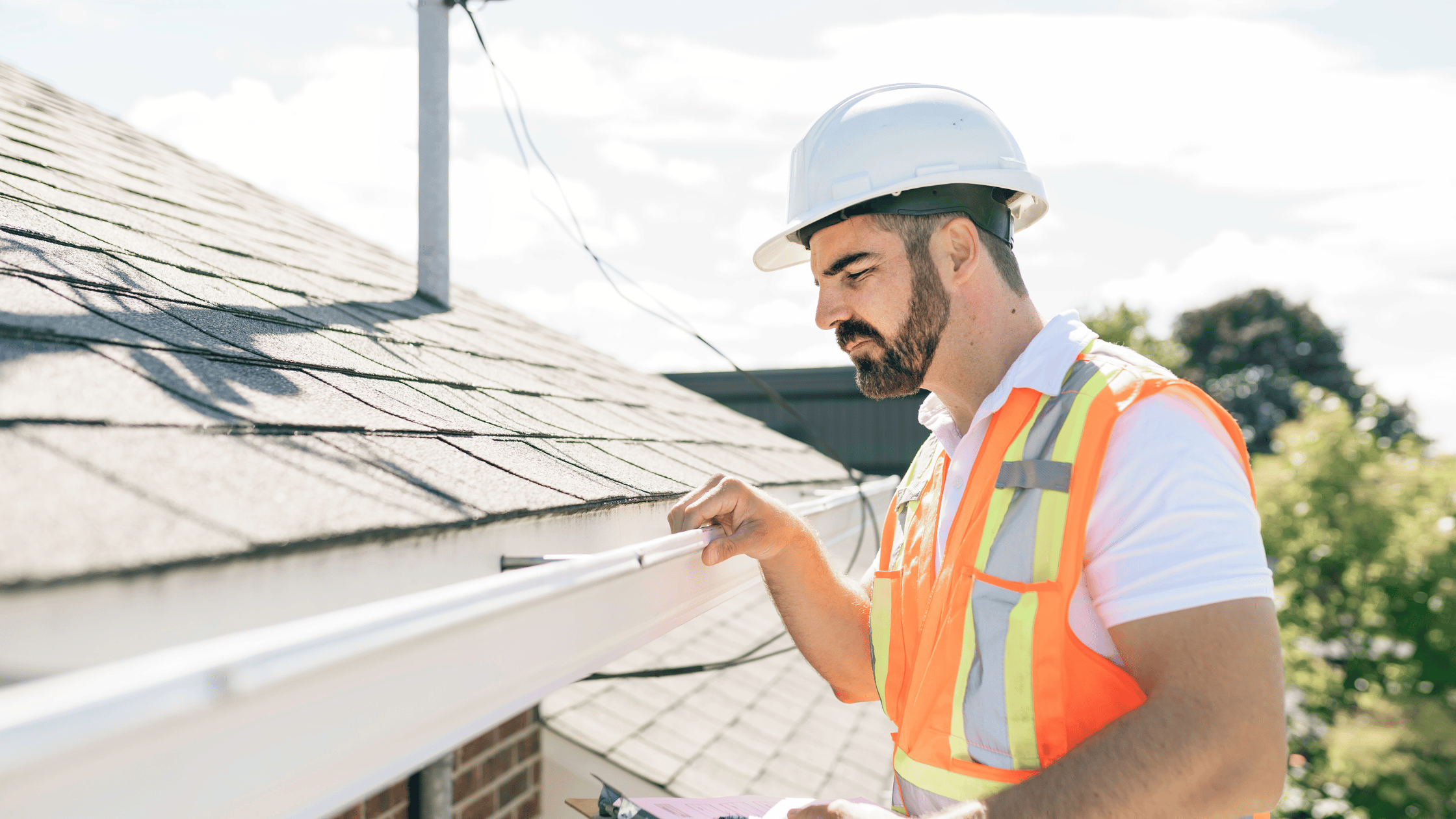 Standing man in a hard hat, holding a clipboard, inspecting home roof